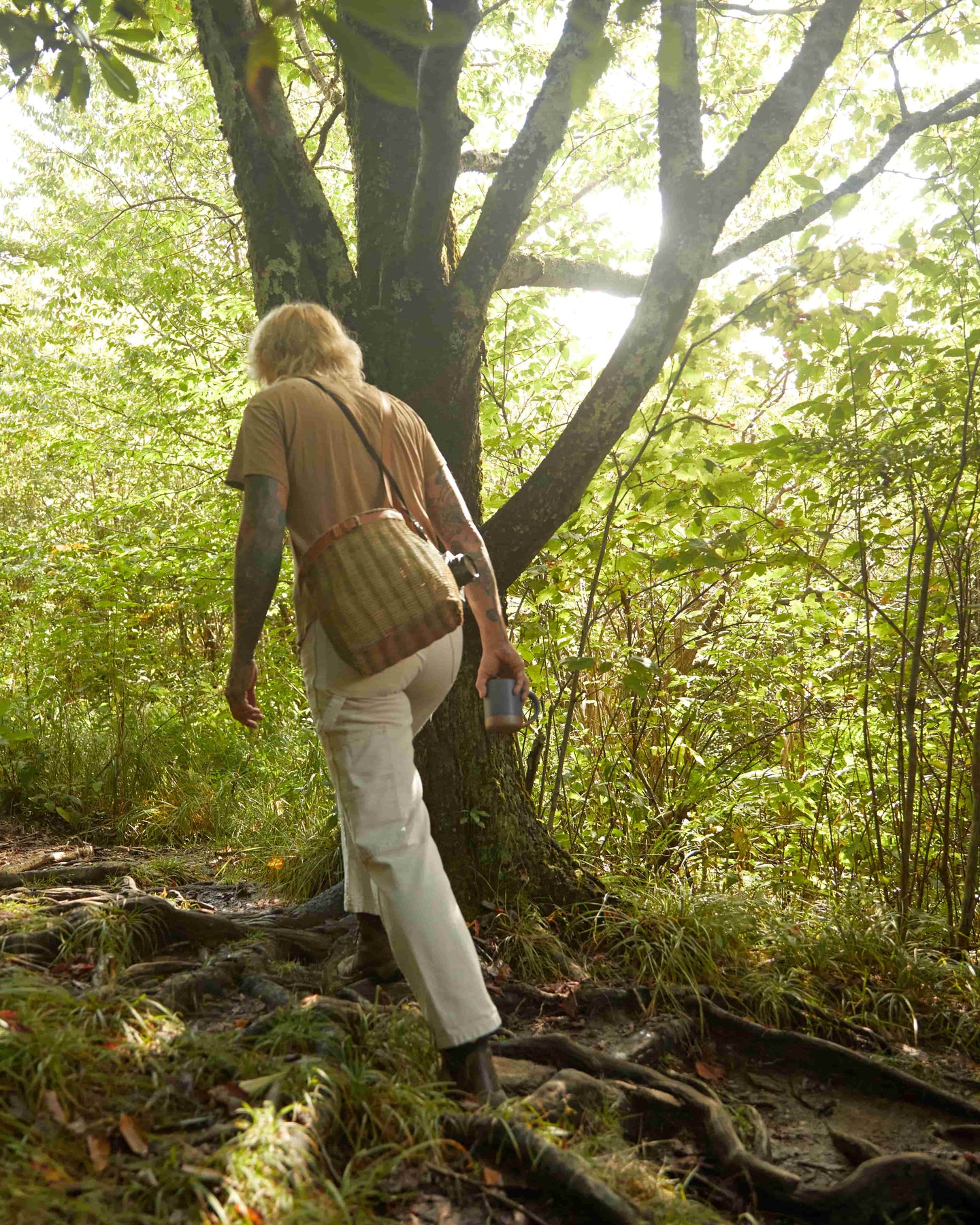 A person walking away in the woods holding a basket bag and carrying a blue mug