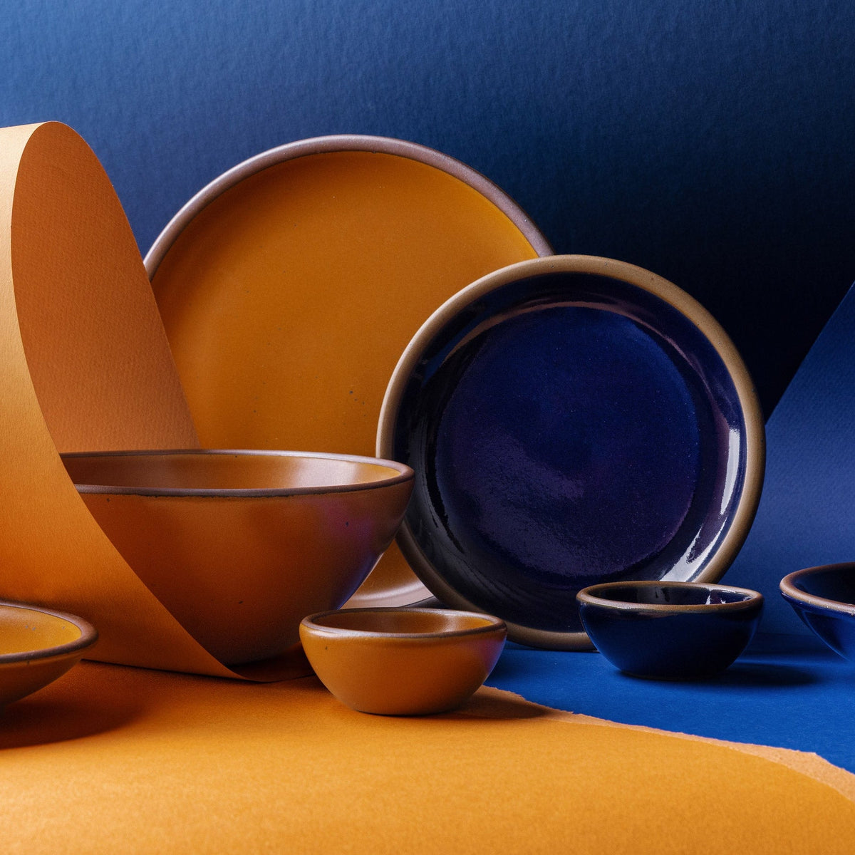 A styled studio arrangement of ceramic tableware in two contrasting glazes: a dark glossy blue and a warm earthy orange. The set includes plate and bowls, set against a split blue and orange paper background.