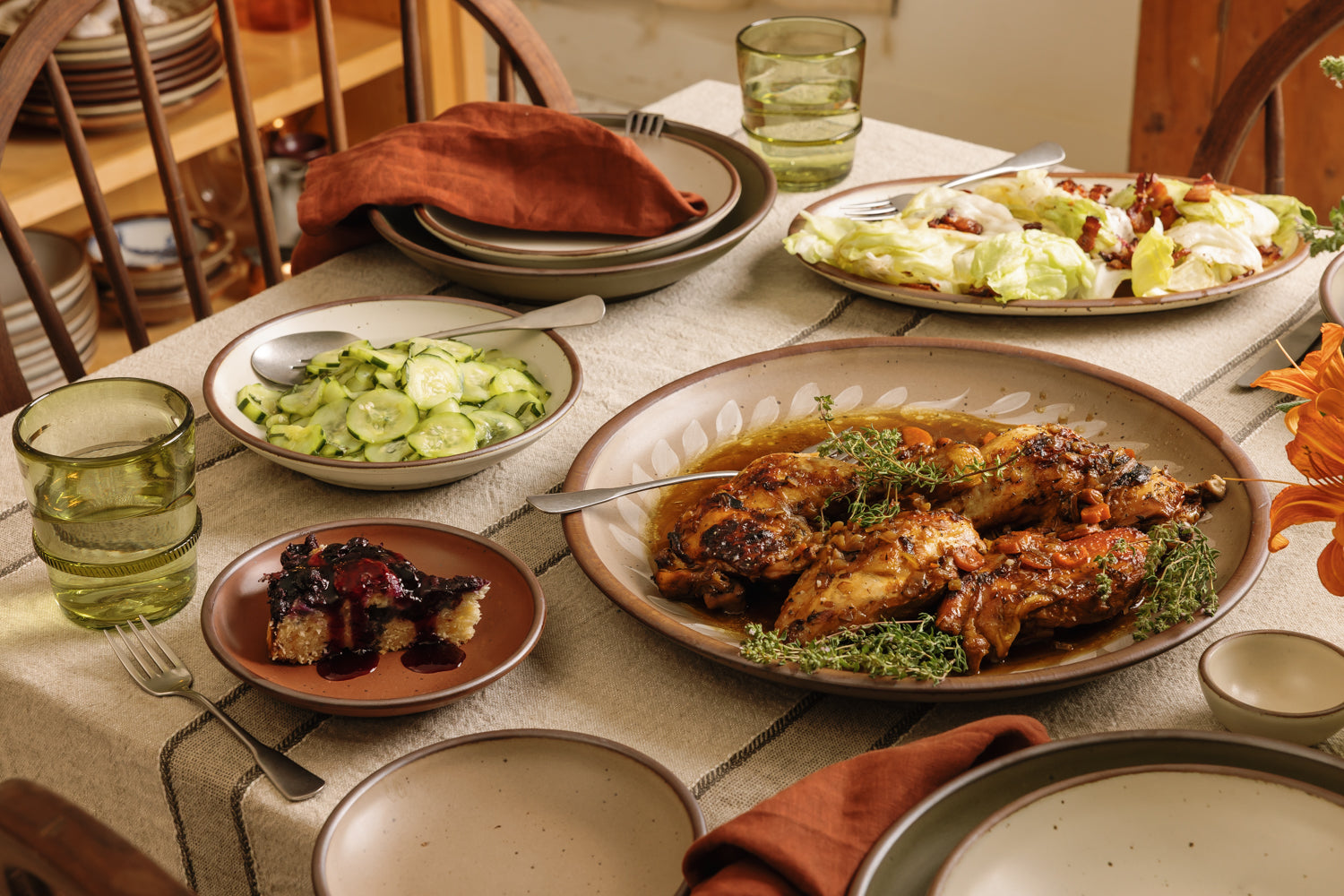 A styled dining table set with multiple dishes, including a large platter of grilled chicken with herbs, a side salad, sliced cucumbers, a dessert with berry sauce, and green-tinted drinking glasses, in a warm indoor setting.