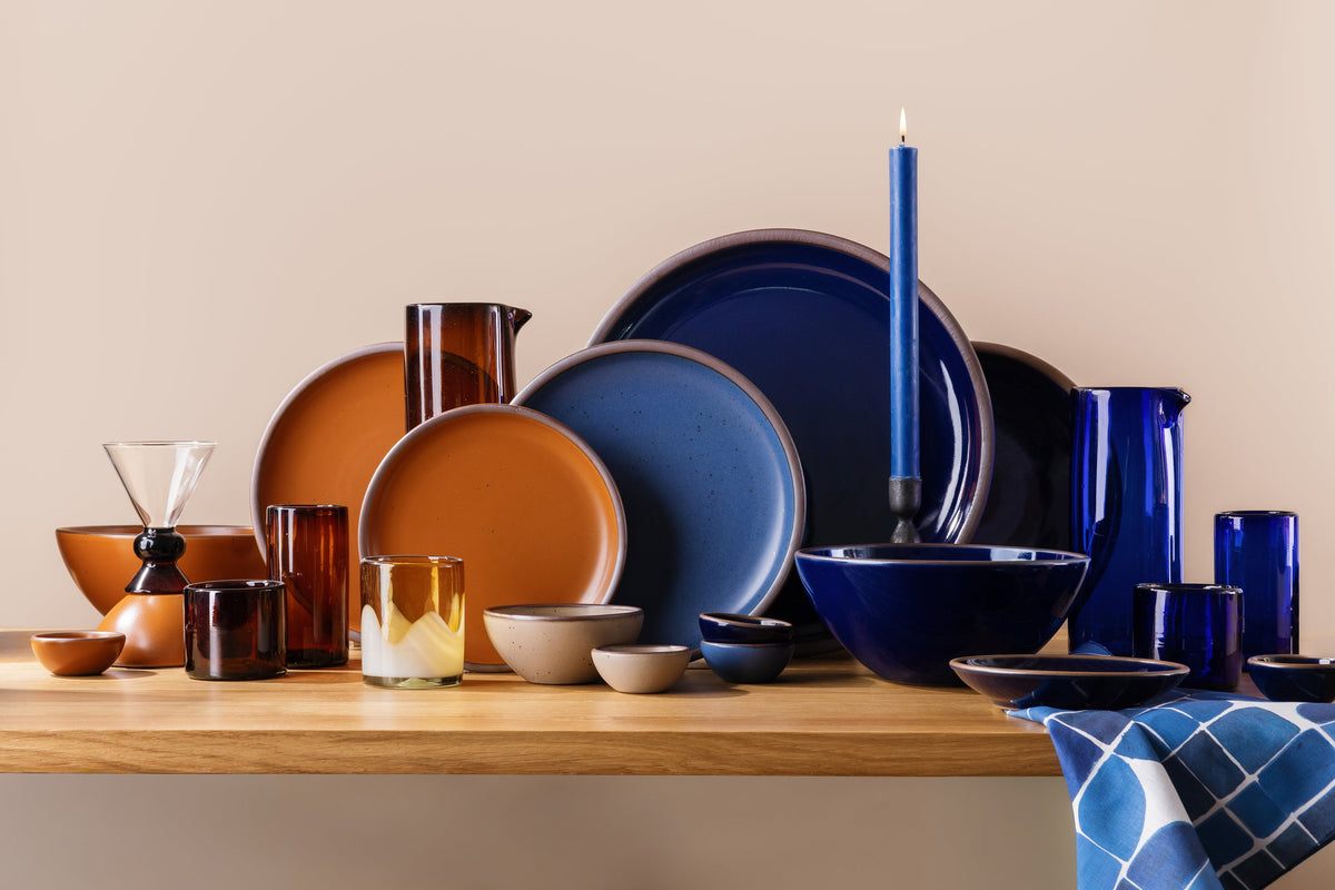 Orange and blue ceramic dinnerware with blue and amber glassware, a lit taper candle, and patterned blue linen.