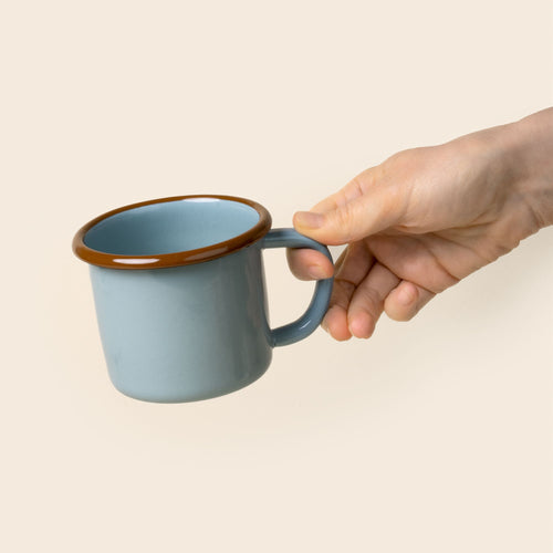 A hand holds out a robin's egg blue enamel mug with a glossy brown rim and handle, shown against a neutral beige background.