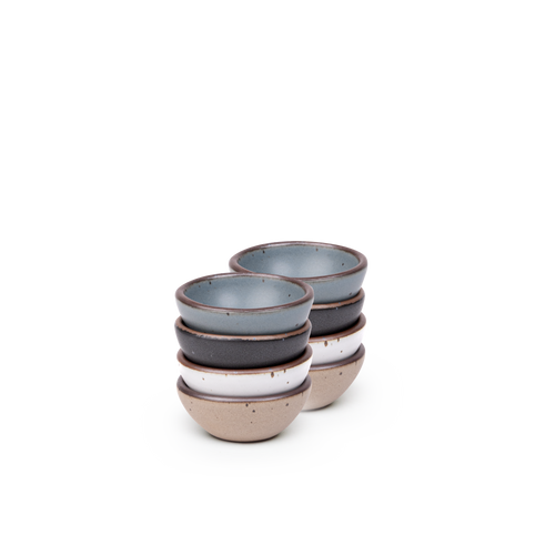 A stack of 8 tiny rounded ceramic bowls in a blue-grey, graphite black, cool white, and warm pale brown colors featuring iron speckles and an unglazed rim