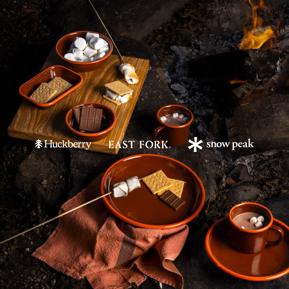 Colorful red enamelware plates, bowls, dishes and mugs around a campfire making s'mores, with overlay text that reads 'Huckberry, East Fork, Snow Peak'
