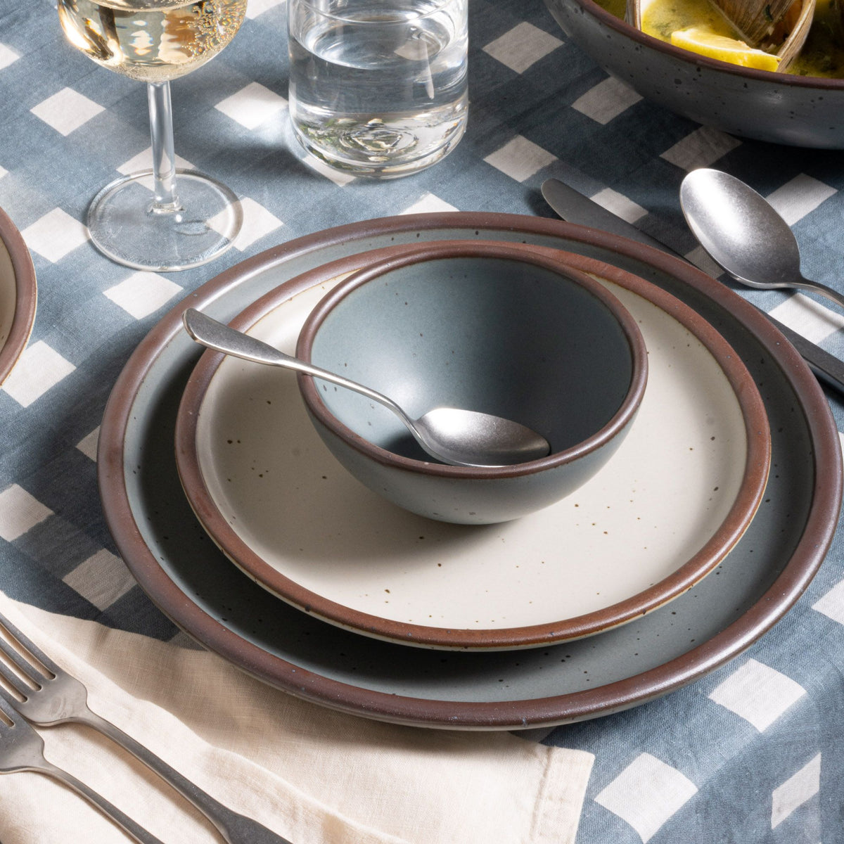 Small round bowl and dinner plate in a blue-grey color with a cream side plate, with silverware on a blue-grey crosshatch tablecloth