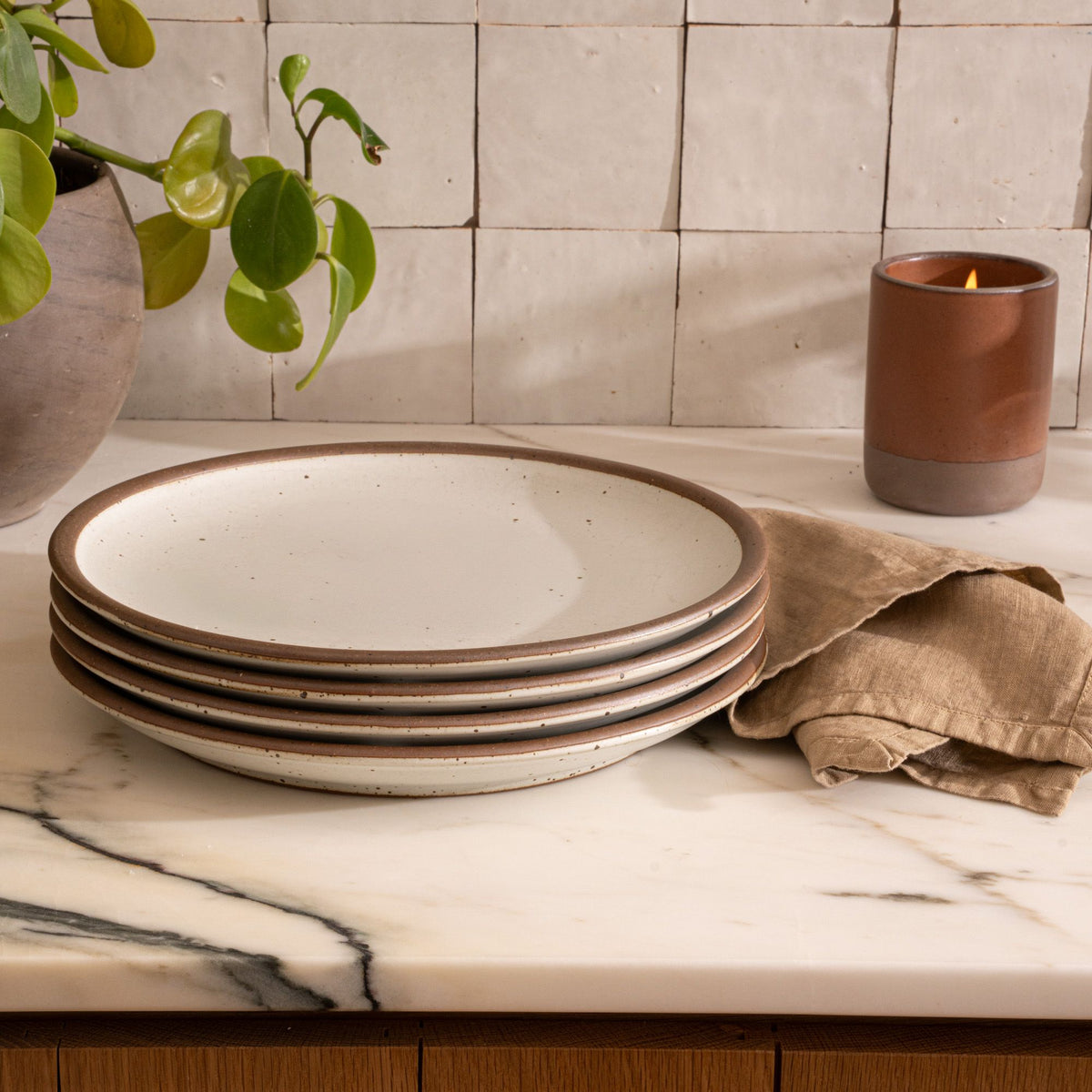 A stack of 4 dinner sized ceramic plates in a cool white color featuring iron speckles and an unglazed rim, with a napkin and lit candle on a countertop