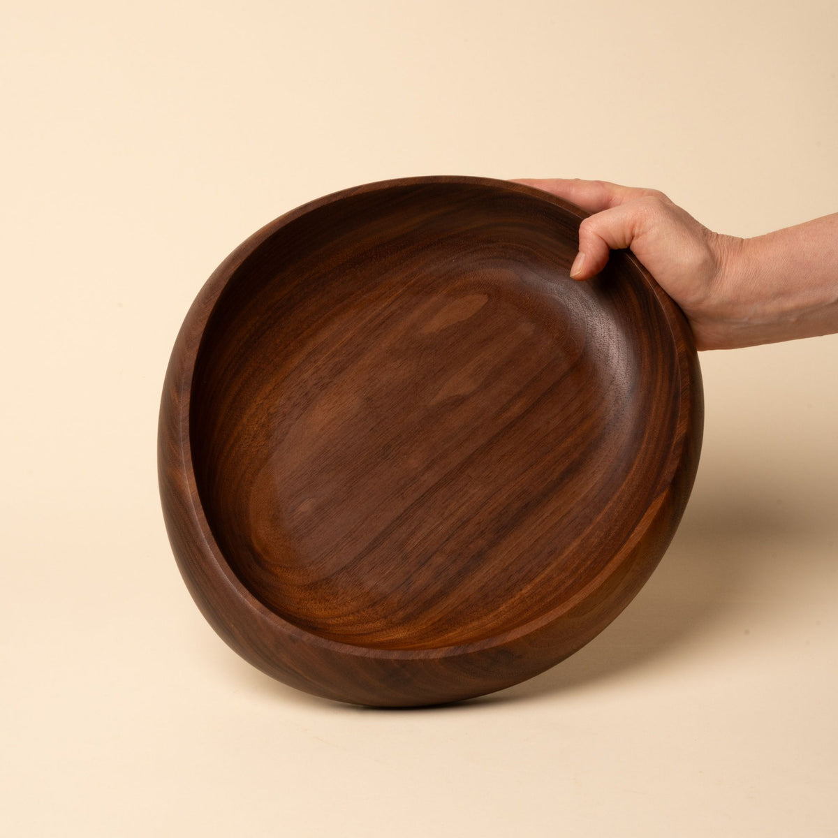 Hand holds out a shallow carved round walnut wood dish with smooth curved edges, resting on a beige background.