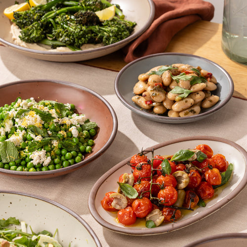 Ceramic coupe bowls and small oval platter with roasted tomatoes, white beans, peas, and broccolini on a linen-covered table.