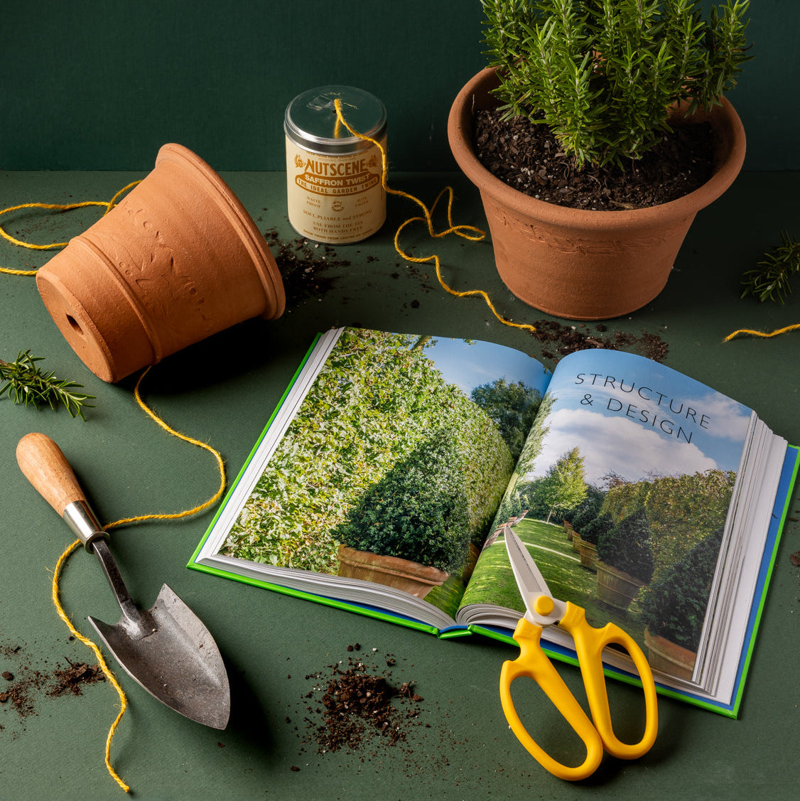 A styled flat-lay of gardening tools and books on a dark green surface. An open book with a page that reads 'STRUCTURE & DESIGN' lies next to yellow-handled pruning scissors, a terracotta pot tipped over, a potted rosemary plant, a small trowel, 