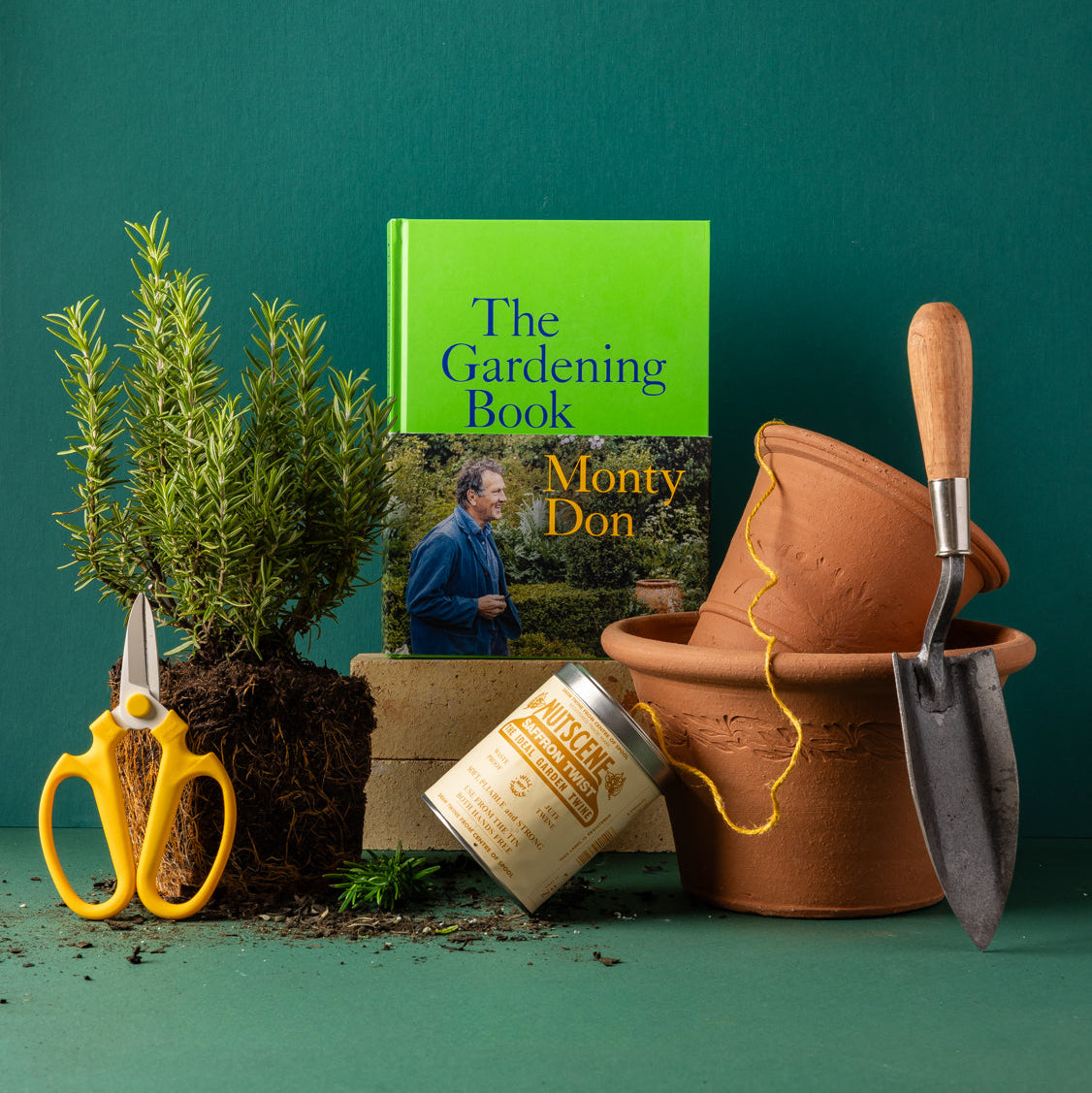 A styled gardening-themed product arrangement on a teal background, featuring a book titled "The Gardening Book" by Monty Don, a small potted rosemary plant, yellow-handled pruning shears, terracotta pots, and a planting trowel.