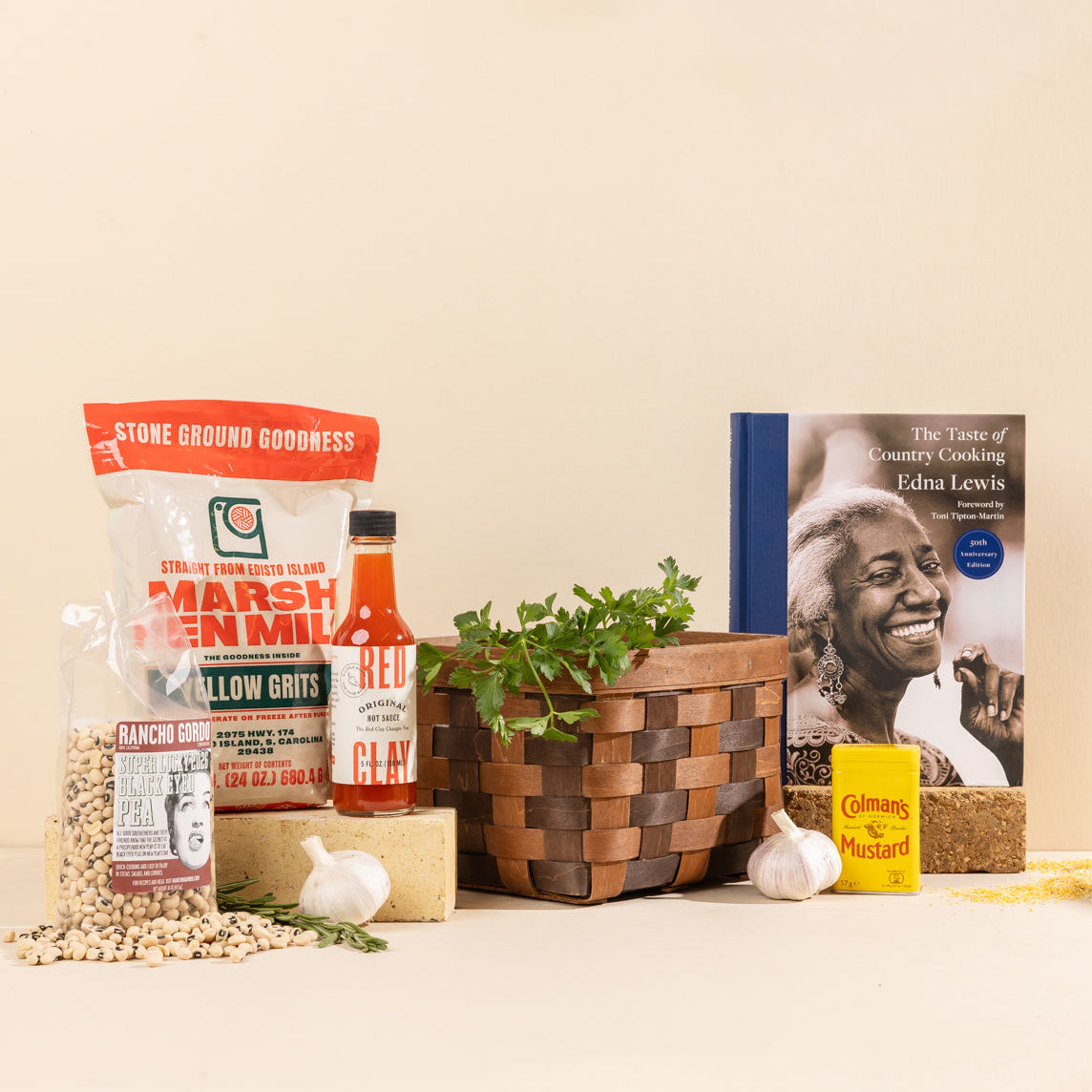 Assortment of Southern pantry staples, including grits, black-eyed peas, hot sauce, mustard, and a The Taste of Country Cooking cookbook arranged around a woven wooden basket.