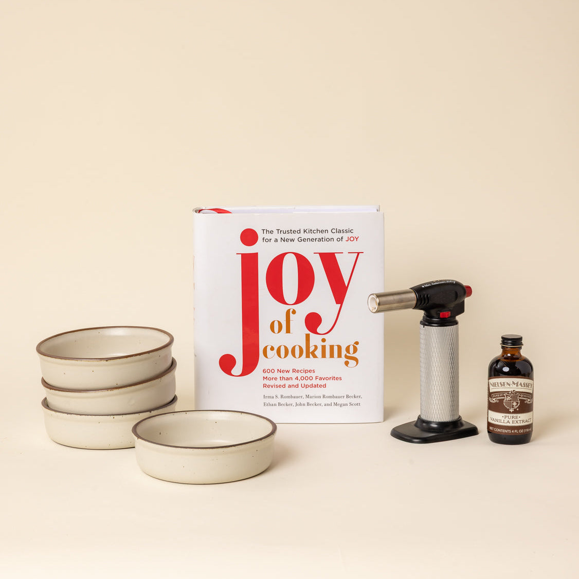 A cookbook titled "Joy of cooking" stands on a warm cream background alongside 4 warm off-white colored, ceramic ramekins, a torch lighter, and a small bottle of vanilla. 