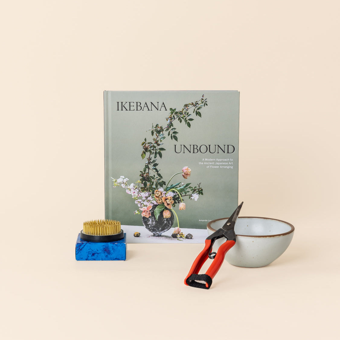 A book titled 'IKEBANA UNBOUND' standing upright, with a medium flower frog on a blue box, red-handled pruning shears, and a small cool white ceramic bowl arranged in front on a warm cream background.