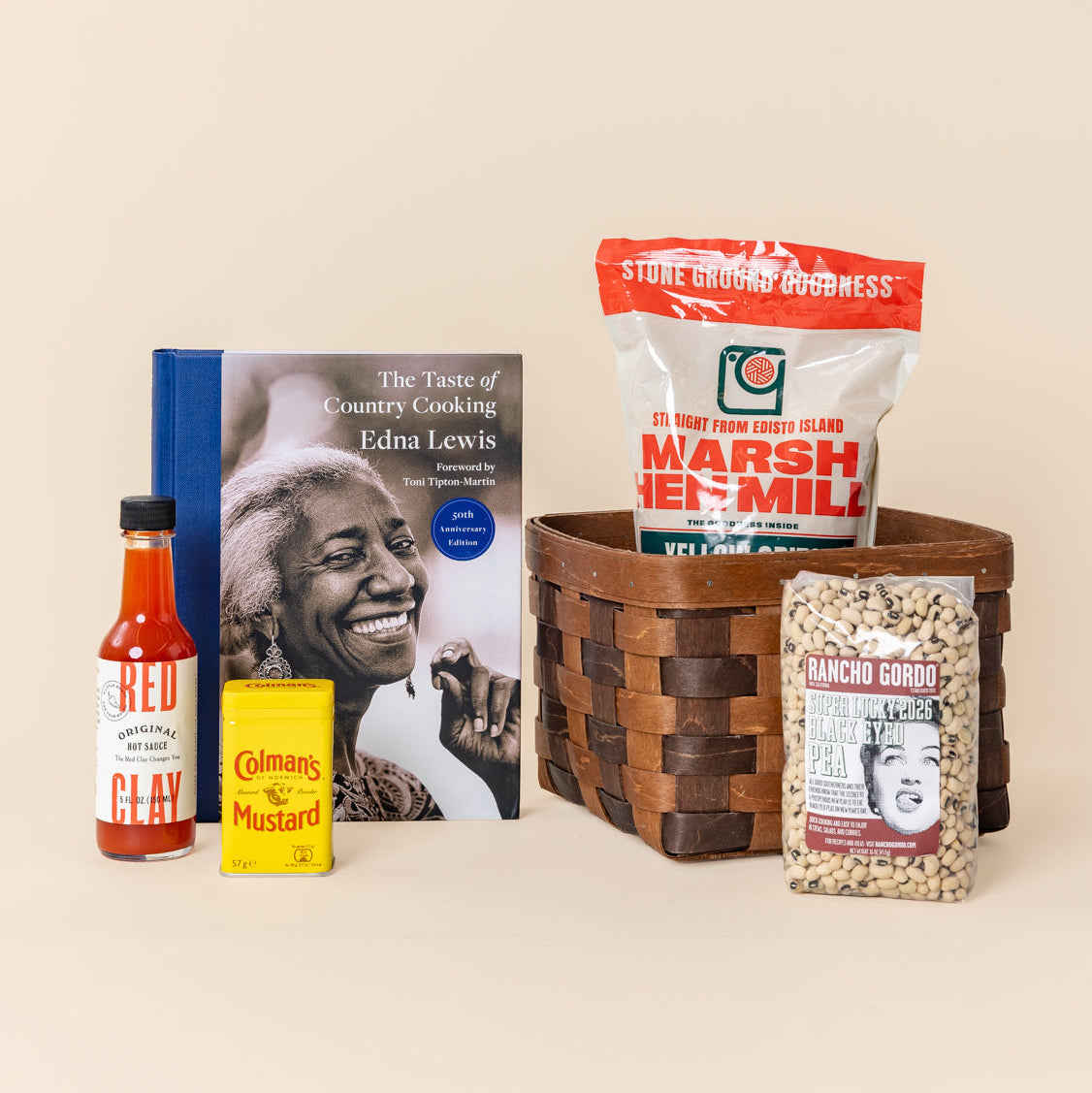 Assortment of Southern pantry staples, including grits, black-eyed peas, hot sauce, mustard, and a The Taste of Country Cooking cookbook arranged around a woven wooden basket.