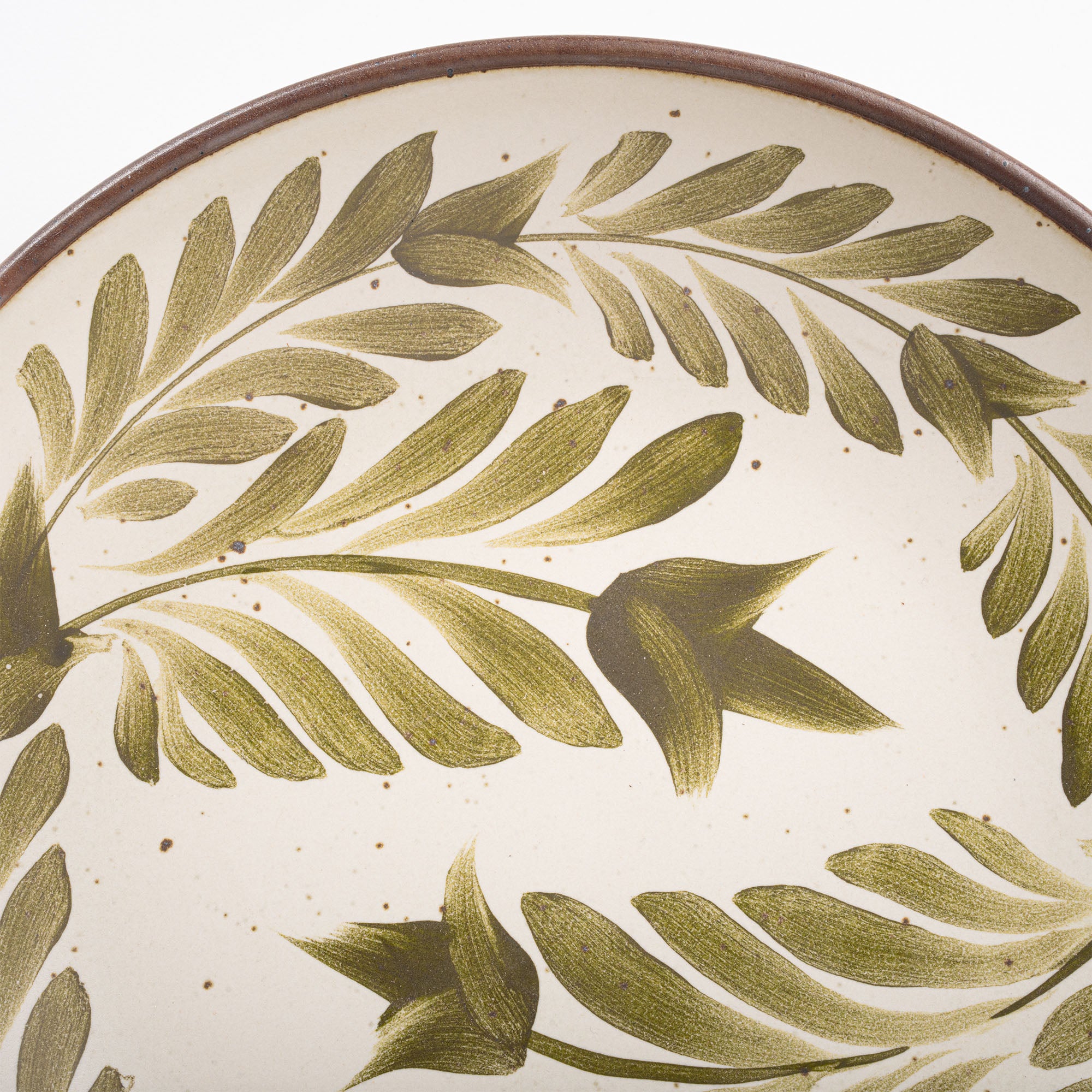A closeup of a large ceramic plate with a curved bowl edge in a warm, tan-toned, off-white color featuring iron speckles and an unglazed rim, with handpainted greenery in an olive green color.