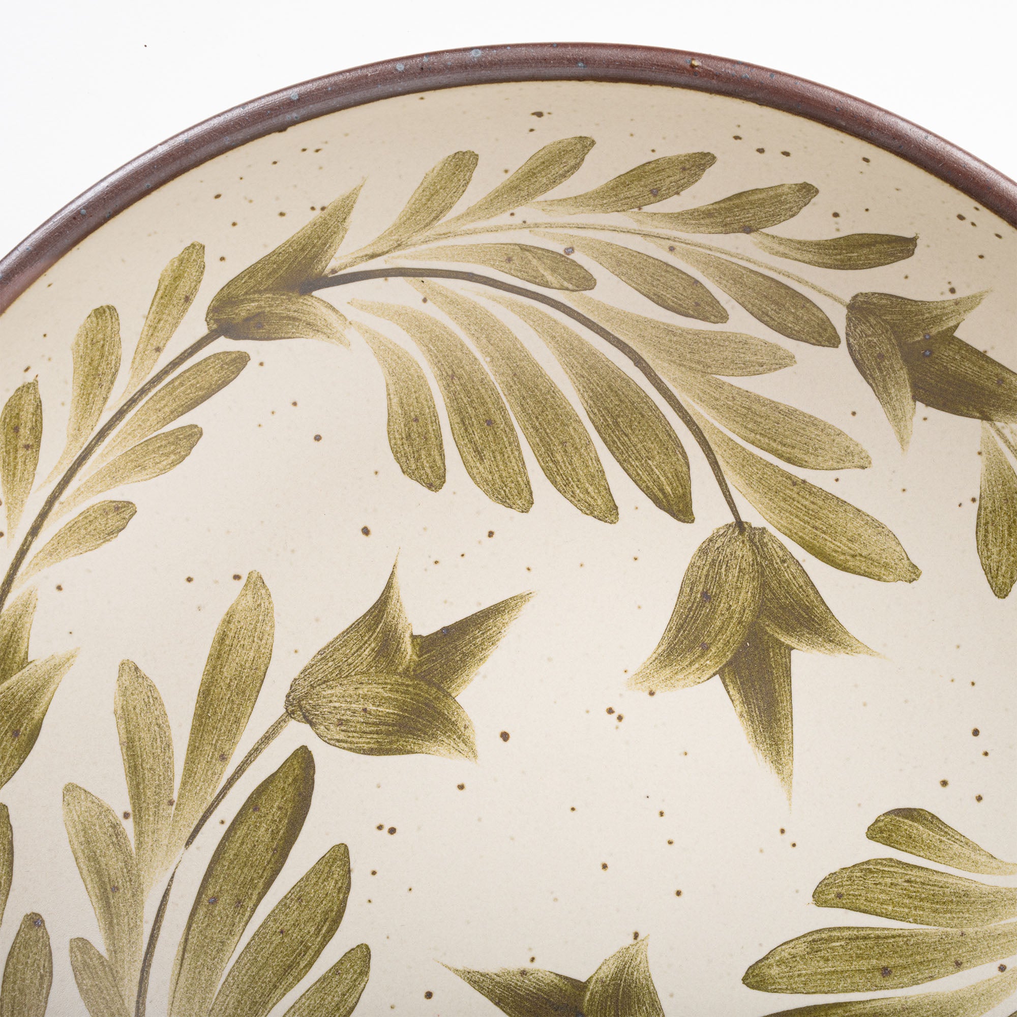 A closeup of a large shallow serving ceramic bowl in a warm, tan-toned, off-white color featuring iron speckles and an unglazed rim, with handpainted greenery in an olive green color inside the bowl.