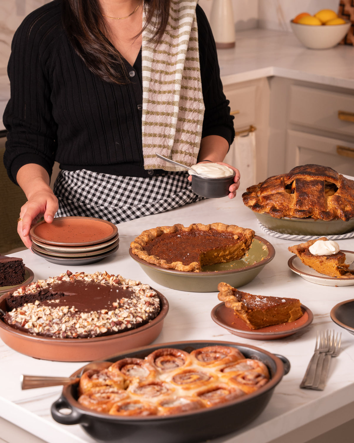 A woman in a kitchen setting presents an assortment of freshly baked desserts, including pumpkin pie, cinnamon rolls, chocolate cake, and pie slices on small plates.