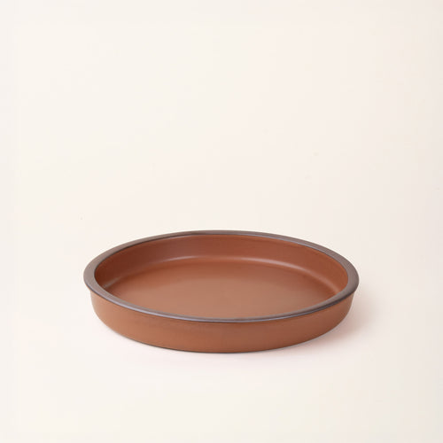 A shallow round ceramic baking dish in a matte terracotta color featuring an unglazed rim, photographed at an angle against a cream background.