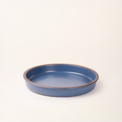 A shallow round ceramic baking dish in a matte grounded blue color featuring an unglazed rim, photographed at an angle against a cream background.
