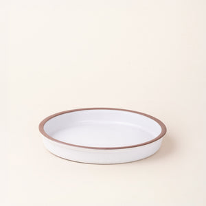 A shallow round ceramic baking dish in a matte cool white color featuring an unglazed rim, photographed at an angle against a cream background.