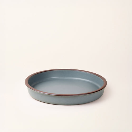 A shallow round ceramic baking dish in a matte blue-grey color featuring an unglazed rim, photographed at an angle against a cream background.