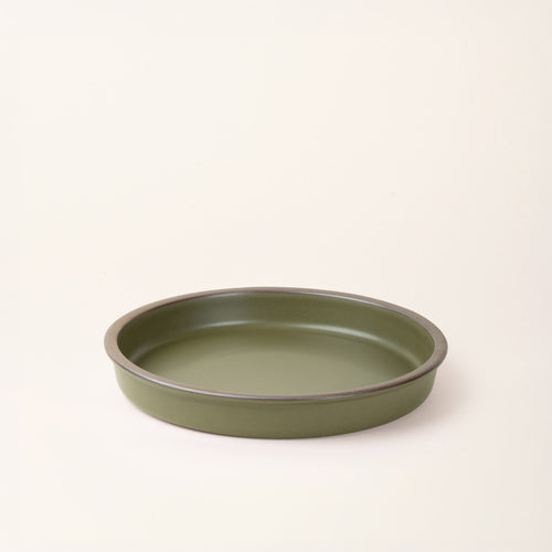 A shallow round ceramic baking dish in a matte olive green color featuring an unglazed rim, photographed at an angle against a cream background.