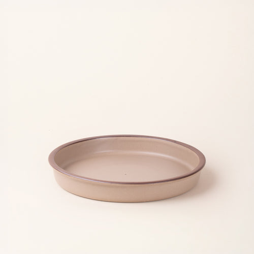 A shallow round ceramic baking dish in a matte pale warm brown color featuring an unglazed rim, photographed at an angle against a cream background.