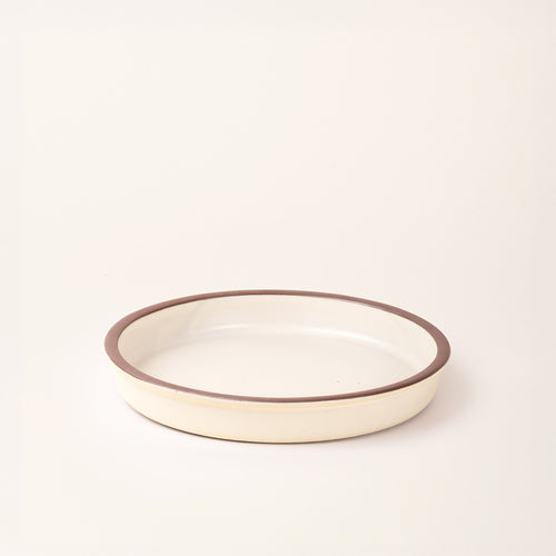 A shallow round ceramic baking dish in a matte warm off-white color featuring an unglazed rim, photographed at an angle against a cream background.