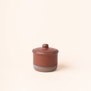 A short ceramic lidded jar in a cool terracotta color with an unglazed base.
