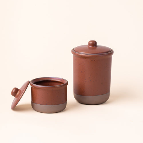 A small and tall ceramic lidded jars in a cool terracotta color with an unglazed base.