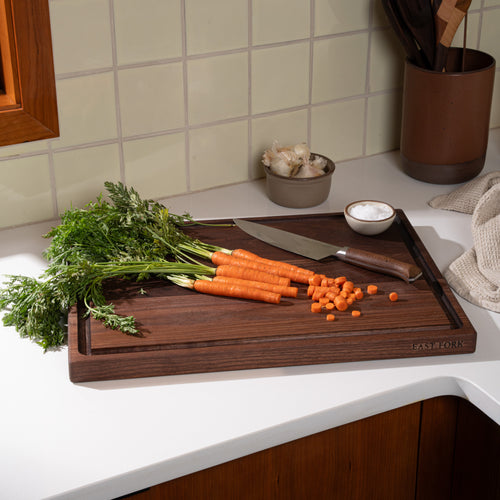 A large walnut cutting board with a juice groove, with carrots mid-cut with a knife with a knife and salt and garlic nearby.