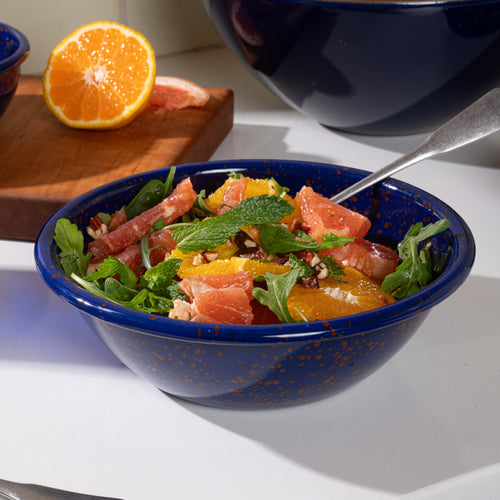 A larger enamel bowl in midnight blue color with red splatter pattern, filled with a fruit salad.