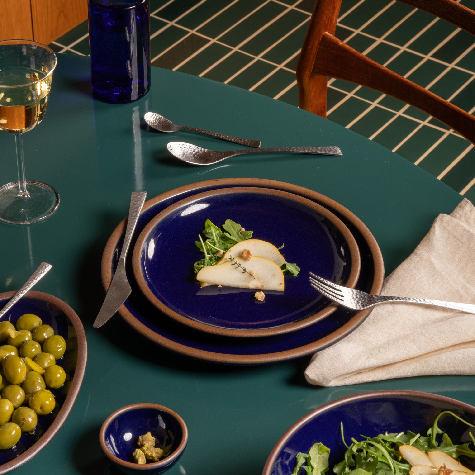 A styled dining table setting featuring a stack of deep glossy blue ceramic plates with a brown rim, silver flatware, a glass of white wine, and side dishes on a teal table.