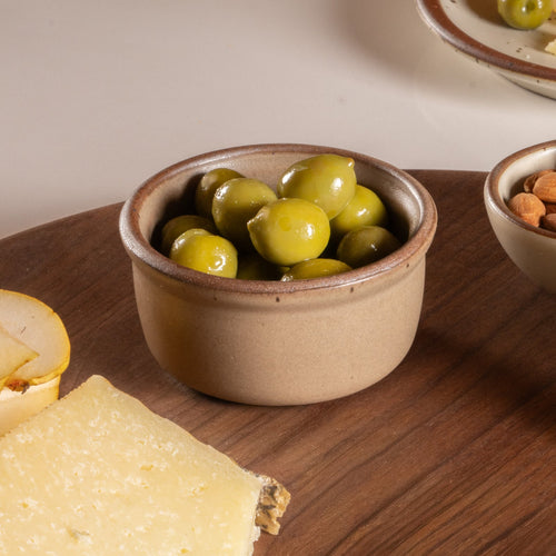Small ceramic ramekin in a warm pale brown color with an unglazed rim, holding olives on a cheeseboard.