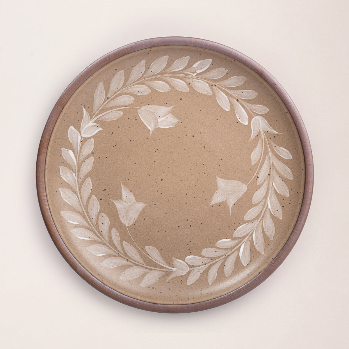Large round ceramic platter with a hand-painted white leaf wreath and floral design on a warm pale brown background with an exposed clay rim.