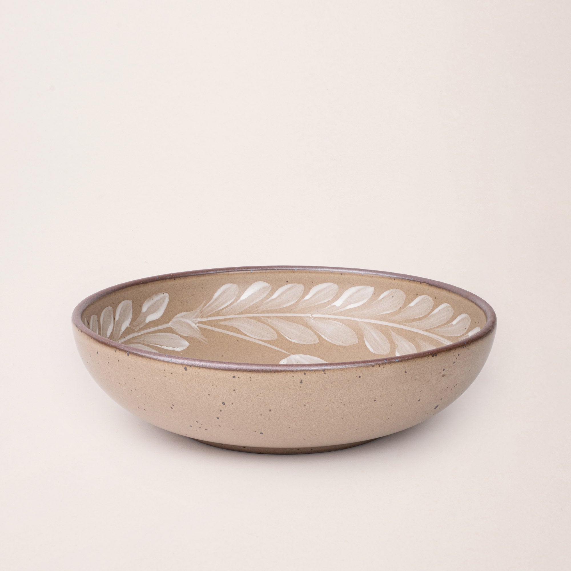 Large ceramic shallow bowl with a hand-painted white leaf border on a pale warm brown interior and exterior with an exposed clay rim.