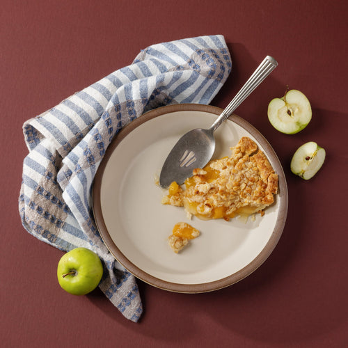 A styled overlay of a shallow round ceramic pie plate in a matte warm off-white color featuring an unglazed rim with a silver pie server, with a slice of pie, along with a striped kitchen towel with blue, tan, and white colors.