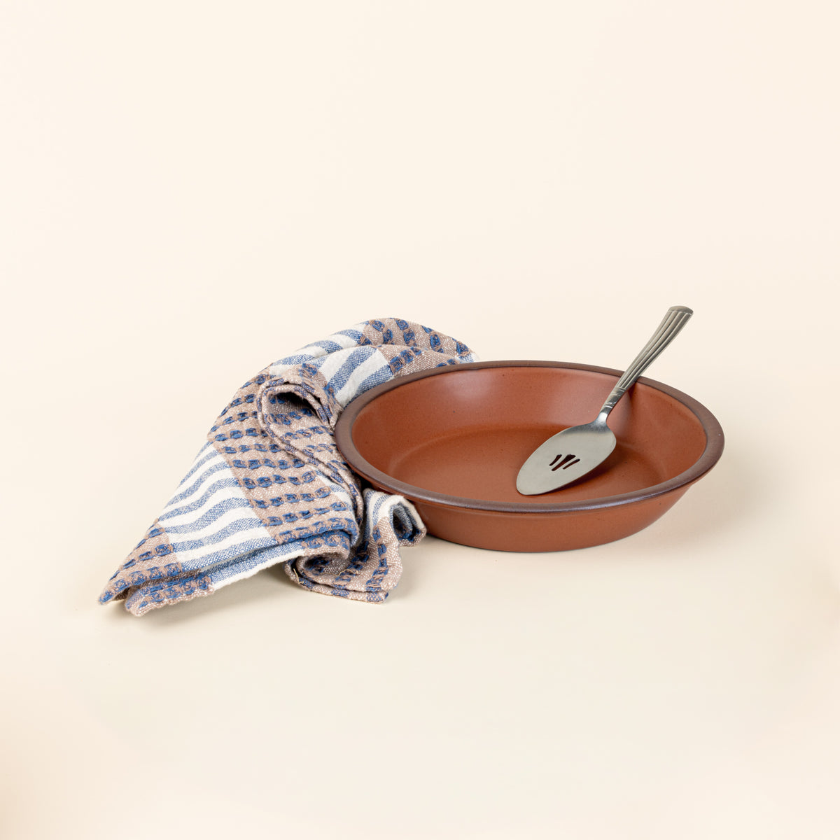 A shallow round ceramic pie plate in a matte terracotta color featuring an unglazed rim with a silver pie server, along with a striped kitchen towel with blue, tan, and white colors.