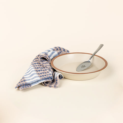 A shallow round ceramic pie plate in a matte warm off-white color featuring an unglazed rim with a silver pie server, along with a striped kitchen towel with blue, tan, and white colors.
