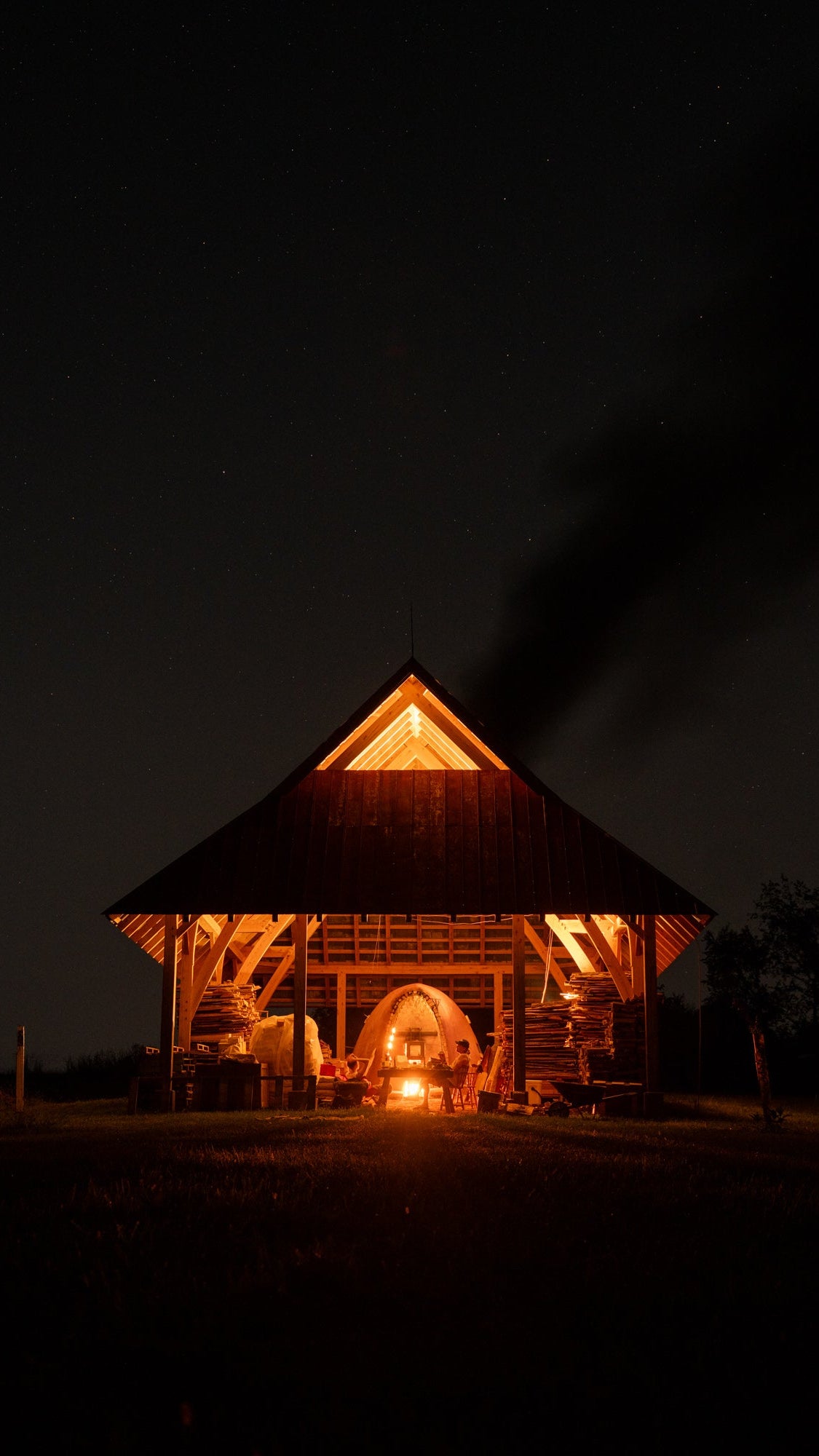 A cabin at night with a wood fired kiln firing with the whole cabin aglow and smoke coming out of the top.