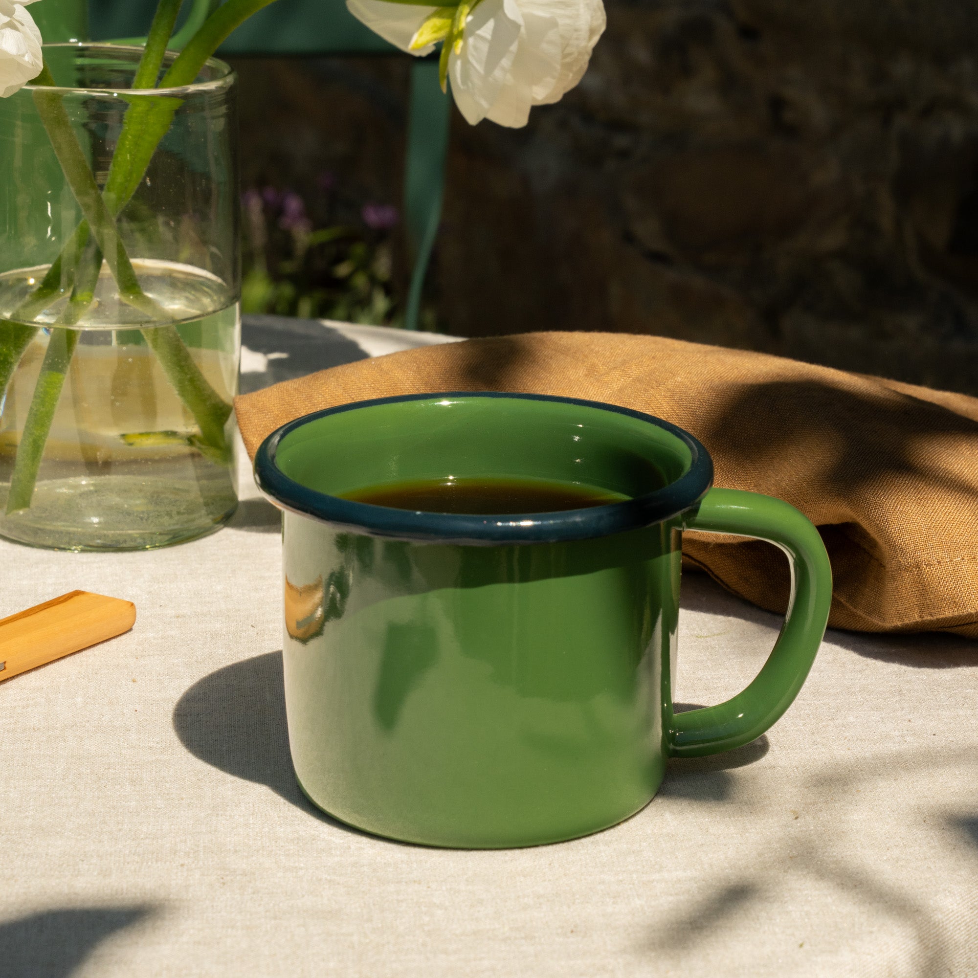 Green enamel mug with dark rim on a light surface, next to a vase with white flowers and a brown napkin.