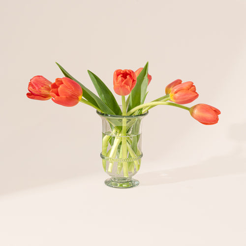 Clear vase with a flared rim, pedestal base, and decorative band detail at the center, filled with flower stems and water.