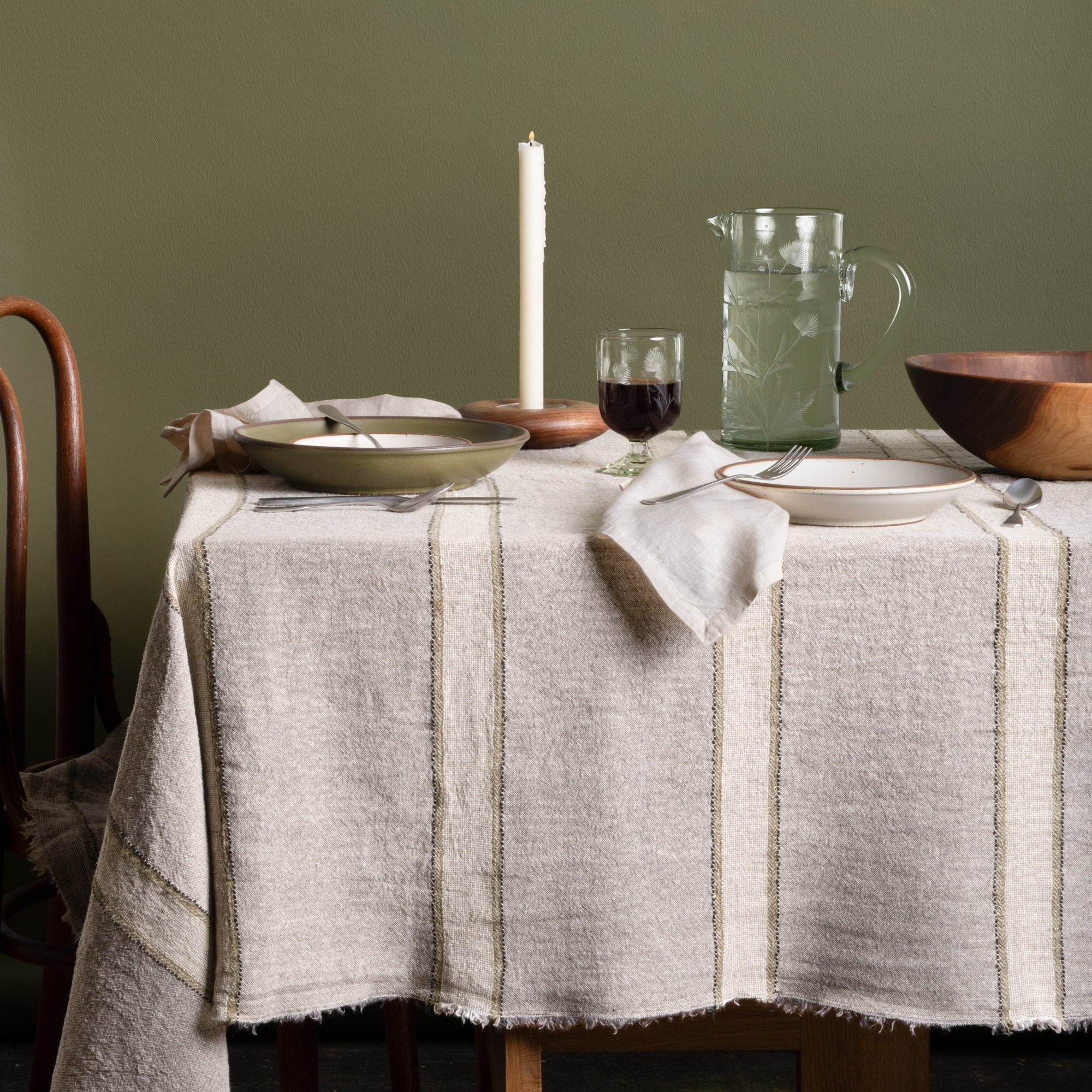Rustic dining table styled with a gray and olive striped linen tablecloth, ceramic dinnerware, glass pitcher, and candlelit accents.