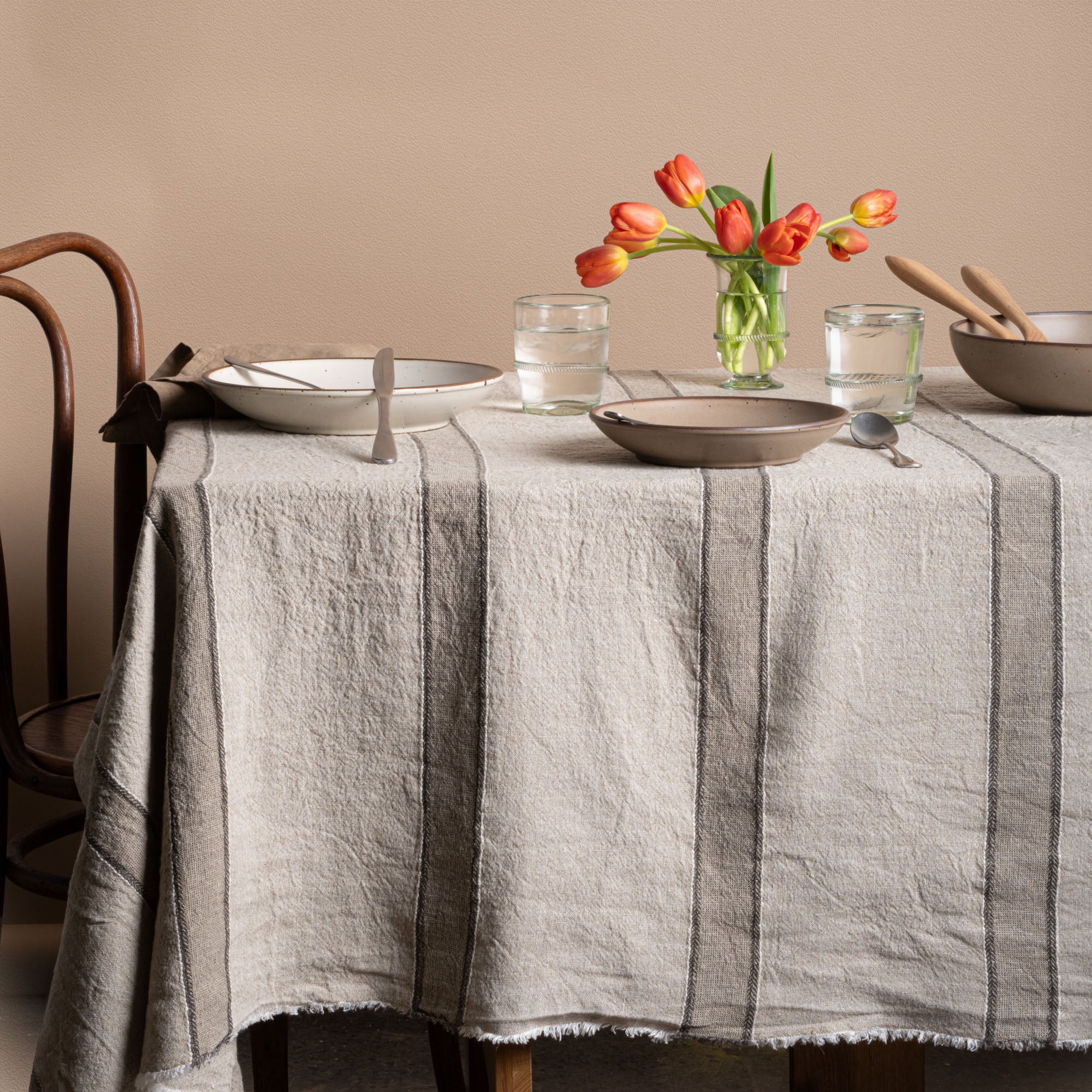 Rustic dining table styled with a neutral striped linen tablecloth, ceramic dinnerware, banded glasses and a vase of tulips.