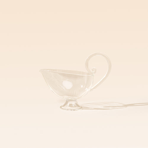 A clear glass sauce boat with a delicate curled handle, pointed pouring spout, and small pedestal foot, photographed on a warm cream background.