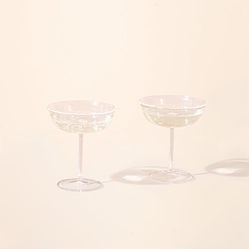 Two clear coupe glasses filled with champagne on a light background.
