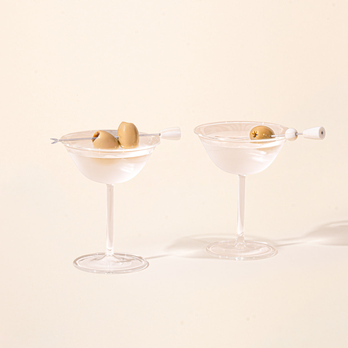 Two clear martini coupe glasses with cocktails, each garnished with green olives on cocktail picks.