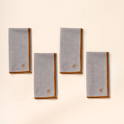 Four folded light blue linen napkins with rust-colored piping and embroidered monograms, arranged on a cream background.