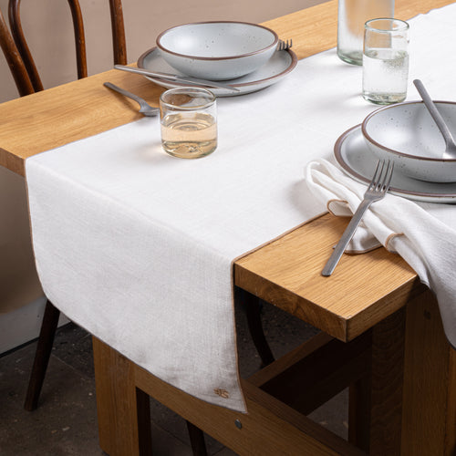 A table set with ceramic pottery, glasses of wine and water, silverware, and a white table linen with taupe-colored piping and embroidered monogram.