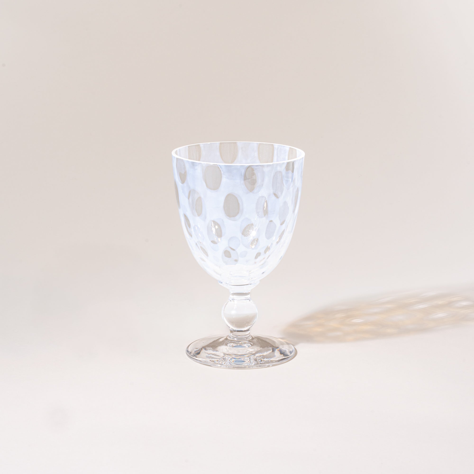A clear stemmed goblet with a subtle polka-dot pattern, photographed empty against a warm neutral background with a soft cast shadow.