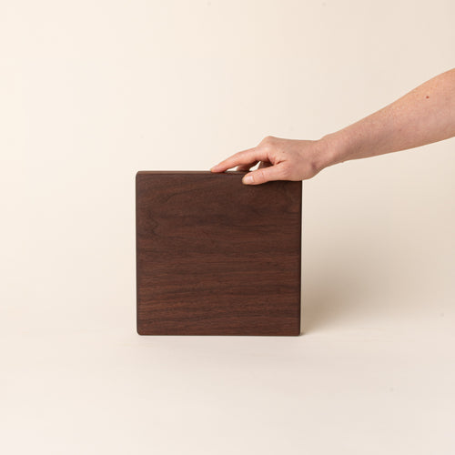 A hand props up a square walnut cutting board on its side